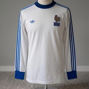 K&ouml;nnte beinhalten: Wei&szlig;es Langarmtrikot mit blauen Akzenten, darunter das Adidas-Logo und das Emblem des franz&ouml;sischen Fu&szlig;ballverbands. Das Trikot hat blaue Streifen auf den &Auml;rmeln sowie einen blauen Kragen und B&uuml;ndchen. Das Trikot wird auf einer Schaufensterpuppe pr&auml;sentiert.