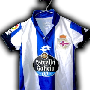 May include: A white and blue soccer jersey with a blue collar and sleeves. The jersey has a vertical blue stripe down the center and features the "Estrella Galicia" logo and a team emblem.