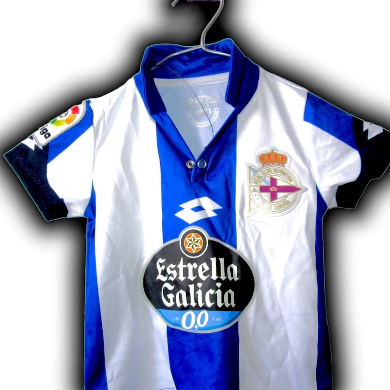 May include: A white and blue soccer jersey with a blue collar and sleeves. The jersey has a vertical blue stripe down the center and features the "Estrella Galicia" logo and a team emblem.