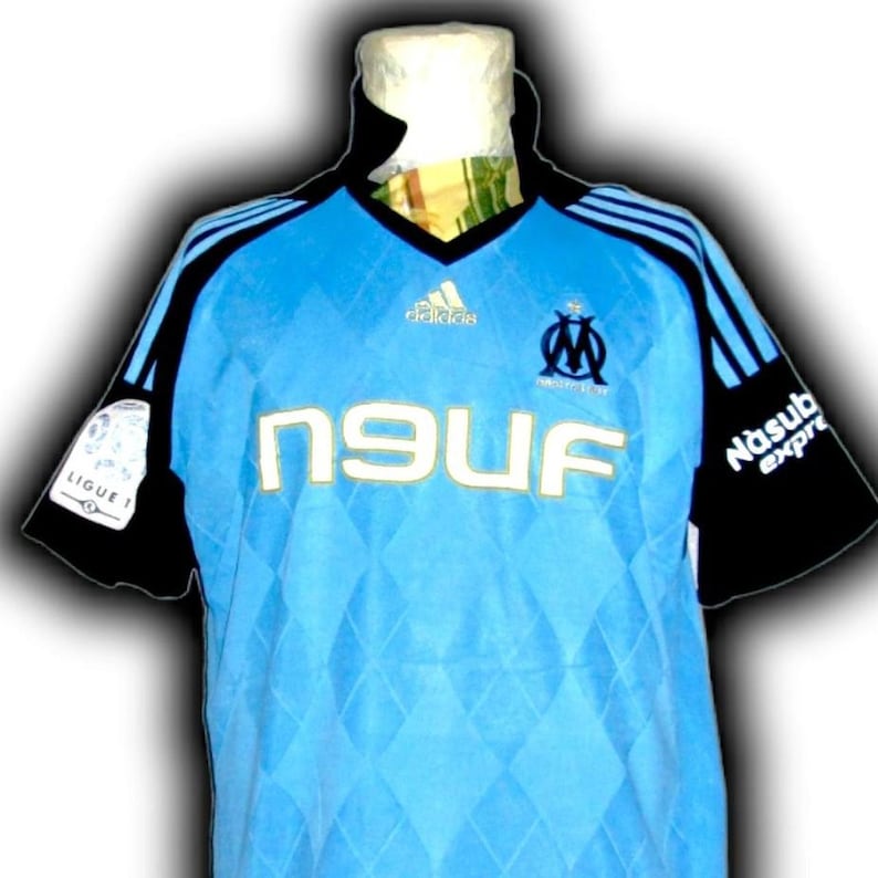 Puede incluir: Camiseta de f&uacute;tbol azul claro con mangas y detalles en negro. La camiseta presenta el logo de Adidas, el emblema del equipo de Marsella y la palabra "NEUF" en letras doradas. Las mangas tienen rayas azules y la camiseta tiene un patr&oacute;n de diamantes.