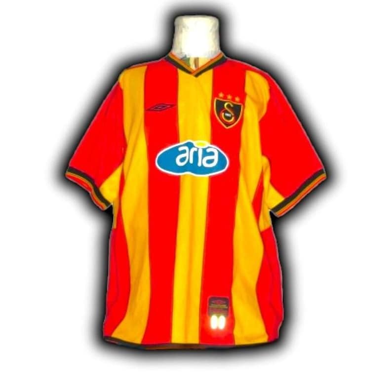 Puede incluir: Una camiseta de f&uacute;tbol a rayas rojas y amarillas con mangas cortas. La camiseta presenta un logotipo ovalado azul con la palabra "aria" en blanco, un escudo del equipo y el logotipo de Umbro. El cuello y los pu&ntilde;os son negros.