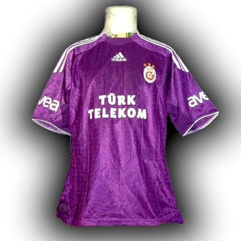 May include: A purple Adidas soccer jersey with white stripes on the shoulders and sleeves. The jersey features the text "T&Uuml;RK TELEKOM" in white, along with the Adidas logo and a team emblem. The sleeves have the text "ave" and "vea".