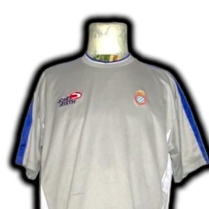 Puede incluir: Una camiseta deportiva gris con detalles azules en las mangas. La camiseta tiene un cuello blanco con ribete azul y presenta el logo "JOHN SMITH" en rojo y un escudo del equipo. La camiseta es de manga corta.