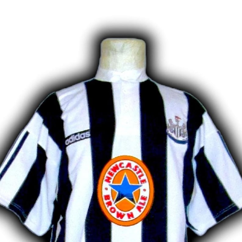 Op de afbeelding: Zwart-wit gestreept voetbalshirt met het Adidas-logo op de schouder. Het shirt heeft een Newcastle Brown Ale-logo en een teamembleem. Het shirt wordt op een mannequin gepresenteerd.