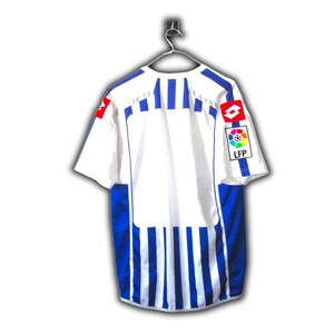 May include: White and blue soccer jersey with a striped design. The jersey has a round neck and short sleeves. Logos are visible on the sleeves. The jersey is hanging on a metal hanger.