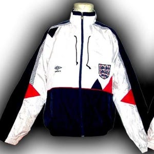 May include: White, navy blue, and black zip-up jacket with red accents. Features the England national team crest and Umbro logo. The jacket has a high collar and a full-length zipper. The sleeves are black, and the body is white and navy blue.