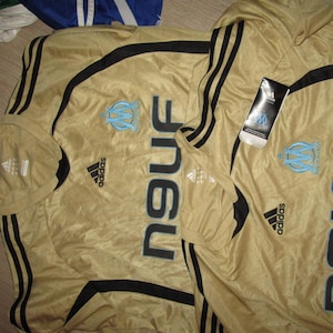 K&ouml;nnte beinhalten: Goldfarbenes Fu&szlig;balltrikot mit schwarzen Streifen auf den &Auml;rmeln und Schultern. Das Trikot zeigt das Teamlogo in Hellblau und das Wort "NEUF" in gro&szlig;en schwarzen Buchstaben. Ein Adidas-Logo ist sichtbar.