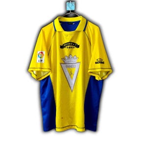 K&ouml;nnte beinhalten: Gelb-blaues Fu&szlig;balltrikot mit dem Text "Ascenso 2003" und "Cadiz C.F.". Das Trikot hat einen Kragen und kurze &Auml;rmel. Das Trikot hat die Logos LFP und Kelme.