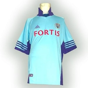 Puede incluir: Camiseta de fútbol azul claro con la palabra "FORTIS" en rojo en el pecho. La camiseta tiene detalles morados en el cuello, las mangas y los laterales. El logo de Adidas es visible, así como un escudo del equipo en el lado derecho.