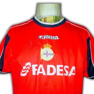 K&ouml;nnte beinhalten: Ein rotes Fu&szlig;balltrikot mit wei&szlig;en und marineblauen Akzenten. Das Trikot zeigt das "Joma"-Logo, ein Teamemblem und das Wort "FADESA" in Wei&szlig;. Der Kragen ist marineblau mit wei&szlig;em Besatz.