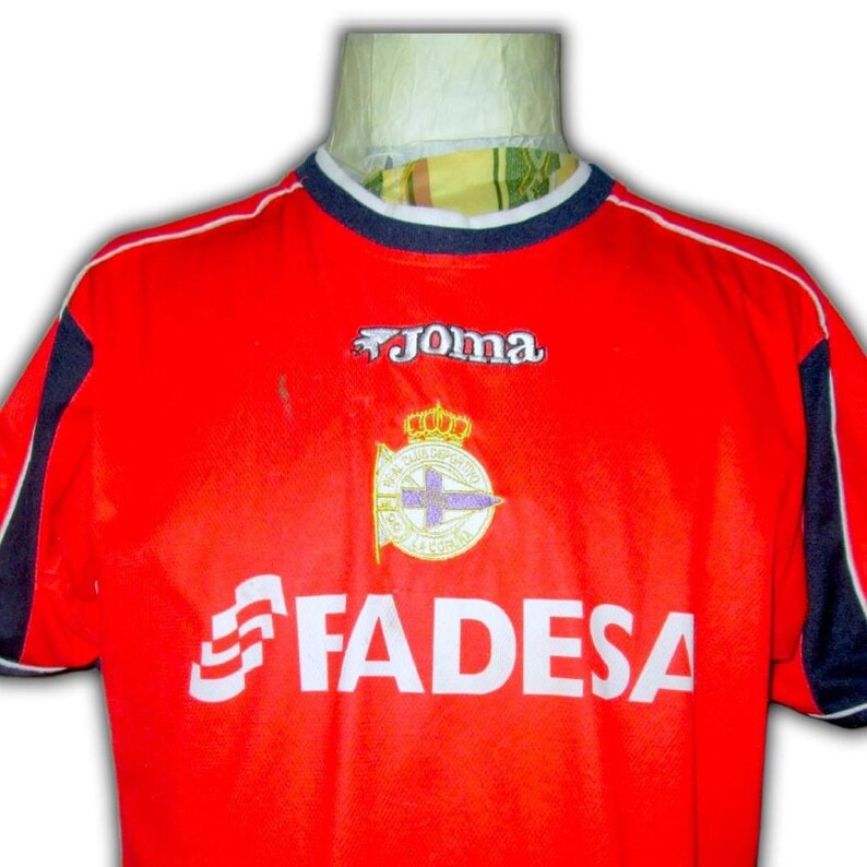 K&ouml;nnte beinhalten: Ein rotes Fu&szlig;balltrikot mit wei&szlig;en und marineblauen Akzenten. Das Trikot zeigt das "Joma"-Logo, ein Teamemblem und das Wort "FADESA" in Wei&szlig;. Der Kragen ist marineblau mit wei&szlig;em Besatz.