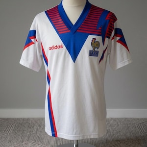 May include: White vintage soccer jersey with blue and red accents. Features a V-neck collar, the Adidas logo, and a French Football Federation emblem. The jersey is displayed on a mannequin.