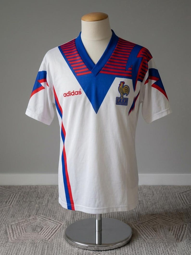 May include: White vintage soccer jersey with blue and red accents. Features a V-neck collar, the Adidas logo, and a French Football Federation emblem. The jersey is displayed on a mannequin.