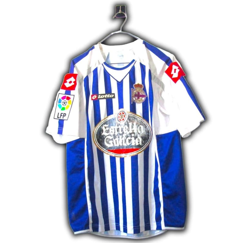 May include: A white and blue striped soccer jersey with a V-neck and short sleeves. The jersey features the "Estrella Galicia" logo, a team crest, and the "Lotto" and "LFP" logos. The jersey is hanging on a metal hanger.