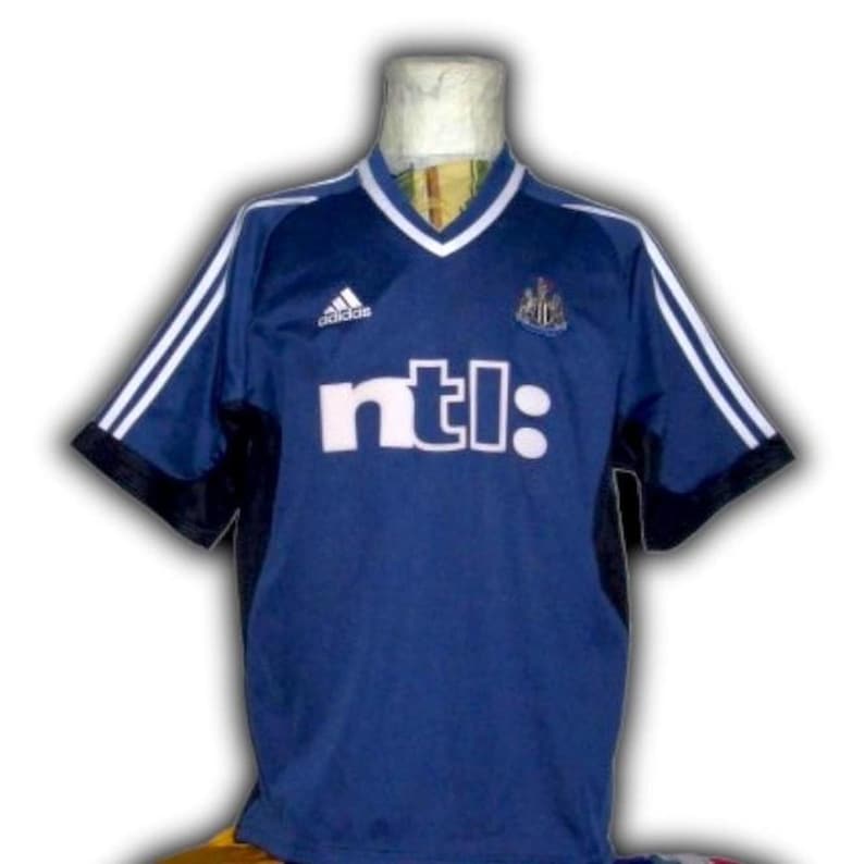 May include: A navy blue soccer jersey with white accents and the text "ntl:" across the front. The jersey has a V-neck collar and white stripes on the sleeves. The Adidas logo is visible on the upper chest.