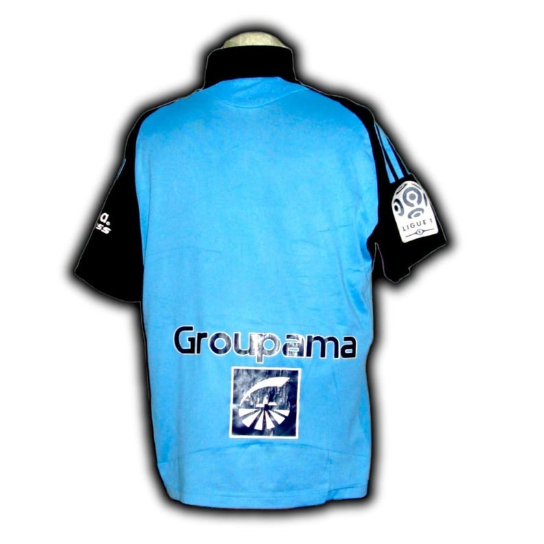 Puede incluir: Camiseta de manga corta azul claro y negra con el texto "Groupama" en la espalda. La camiseta tiene cuello negro y mangas negras. El logo de la Ligue 1 est&aacute; en la manga derecha.