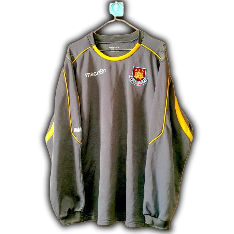 Puede incluir: Camiseta deportiva gris de manga larga con ribetes amarillos y un logotipo del equipo. El logotipo de Macron est&aacute; en el pecho, con "HAM" en la manga. El cuello es amarillo.