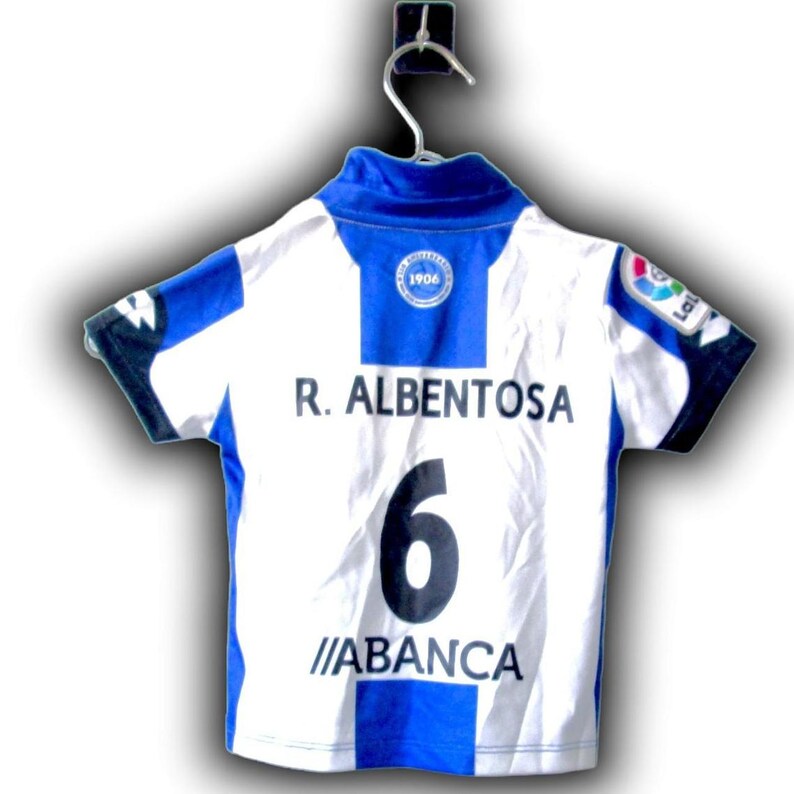May include: A white and blue soccer jersey with the name "R. ALBERTOSA" and the number "6" on the back. The jersey also features the text "ABANCA" and a team logo.