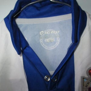May include: A white and blue soccer jersey with a blue collar and accents. The jersey has a logo on the chest and the number 9 on the tag. The jersey is hanging on a metal hanger.