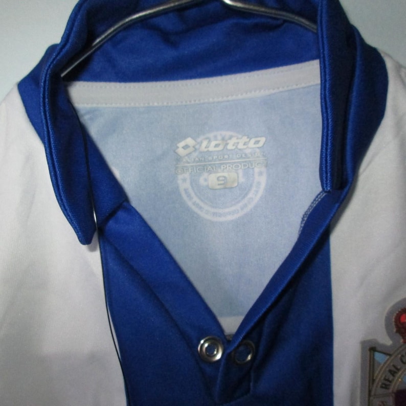 May include: A white and blue soccer jersey with a blue collar and accents. The jersey has a logo on the chest and the number 9 on the tag. The jersey is hanging on a metal hanger.