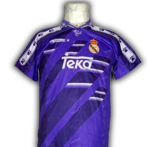 K&ouml;nnte beinhalten: Ein lila-wei&szlig;es Fu&szlig;balltrikot mit dem Teka-Logo und einem Teamwappen. Das Trikot hat diagonale Streifen und Pfotenabdruck-Akzente auf den &Auml;rmeln und am Kragen. Das Kelme-Logo ist ebenfalls sichtbar.