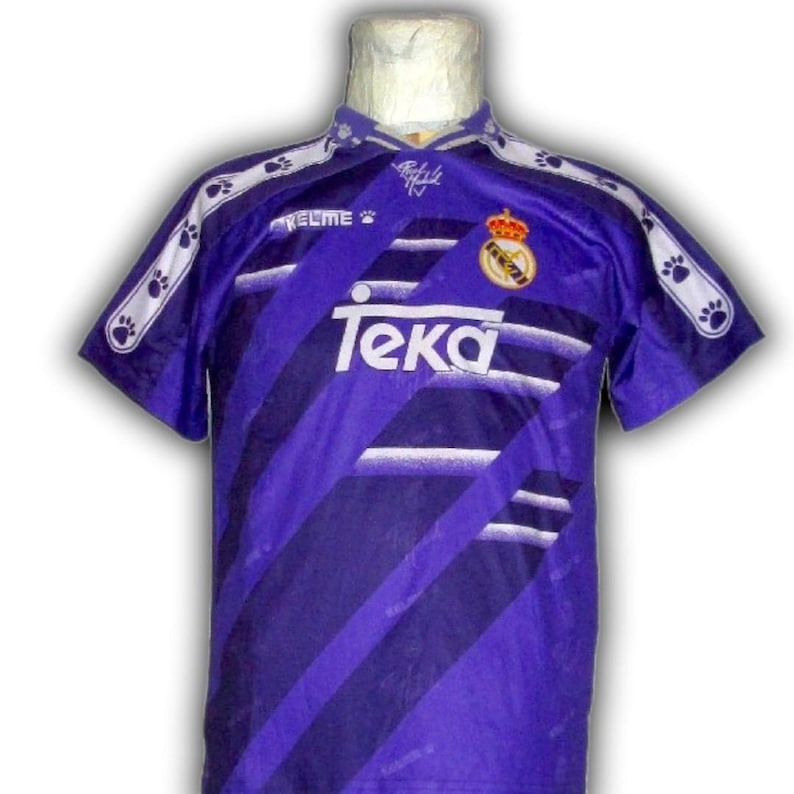 K&ouml;nnte beinhalten: Ein lila-wei&szlig;es Fu&szlig;balltrikot mit dem Teka-Logo und einem Teamwappen. Das Trikot hat diagonale Streifen und Pfotenabdruck-Akzente auf den &Auml;rmeln und am Kragen. Das Kelme-Logo ist ebenfalls sichtbar.