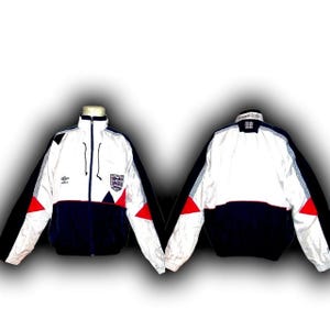 May include: White, navy blue, and red zip-up jacket with the England national team crest. The jacket has a high collar, black accents, and a red triangle design on the sleeves. The Umbro logo is visible on the chest.