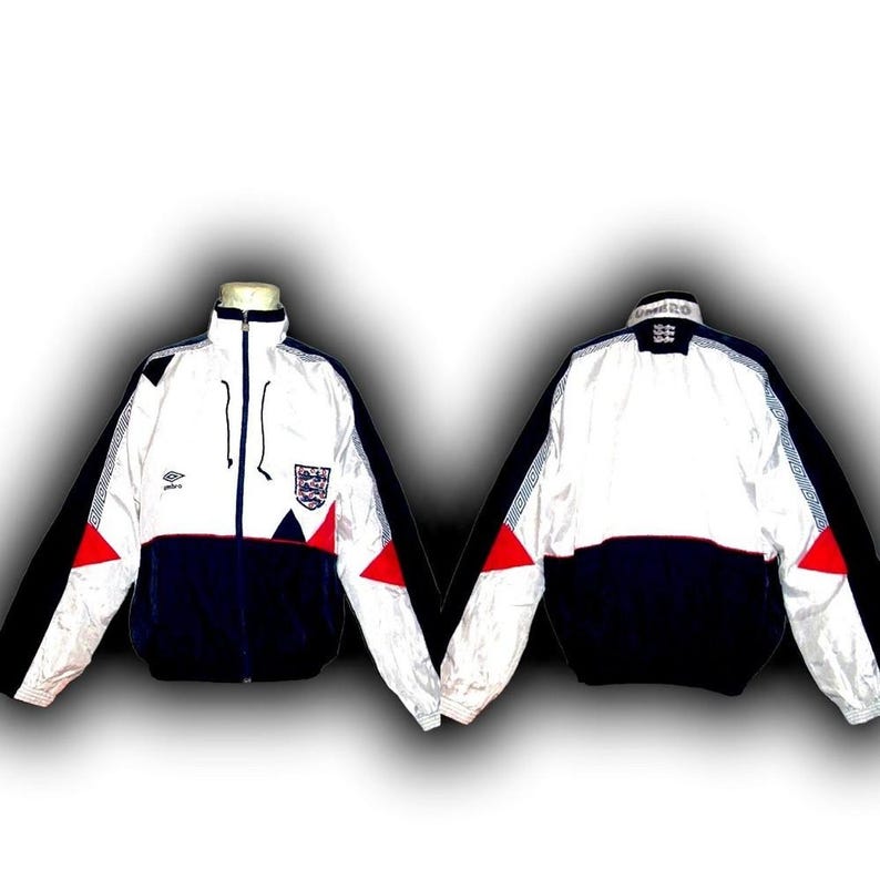 May include: White, navy blue, and red zip-up jacket with the England national team crest. The jacket has a high collar, black accents, and a red triangle design on the sleeves. The Umbro logo is visible on the chest.