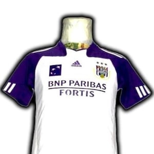 Puede incluir: Camiseta de fútbol blanca y morada con el texto "BNP PARIBAS FORTIS". La camiseta tiene mangas y detalles morados, el logo de Adidas y un escudo del equipo. La camiseta se muestra en un maniquí.