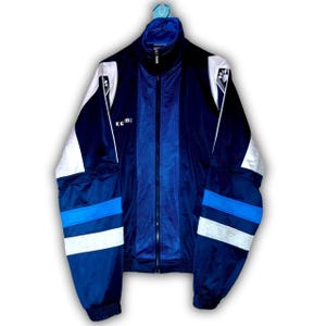 K&ouml;nnte beinhalten: Marineblaue Trainingsjacke mit Rei&szlig;verschluss und wei&szlig;en und hellblauen Akzenten. Die Jacke hat ein Mesh-Panel in der Mitte und den Markennamen "KEM" auf der Brust. Die &Auml;rmel haben wei&szlig;e und hellblaue Streifen.