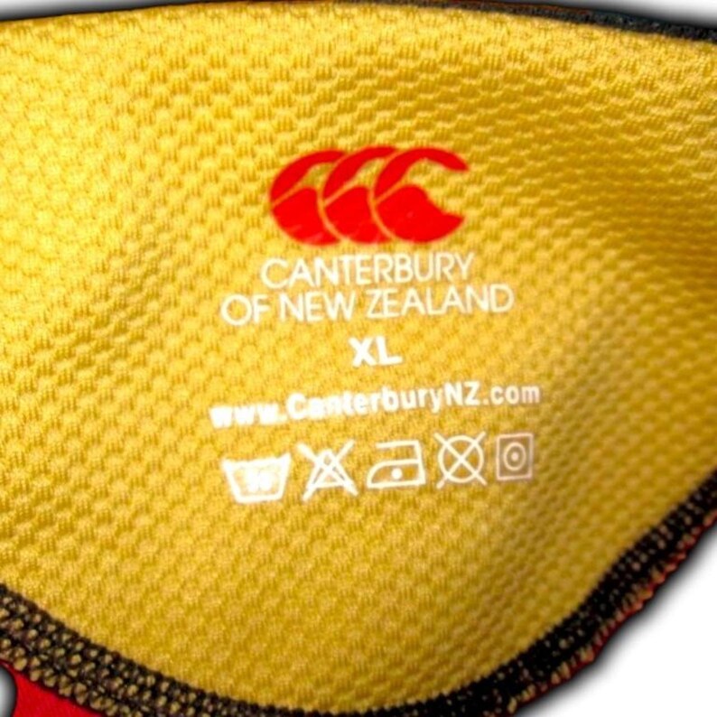 May include: Close-up of a yellow fabric with the Canterbury of New Zealand logo in red. The text "Canterbury of New Zealand" and "XL" are printed in white. The website address www.CanterburyNZ.com is also visible.