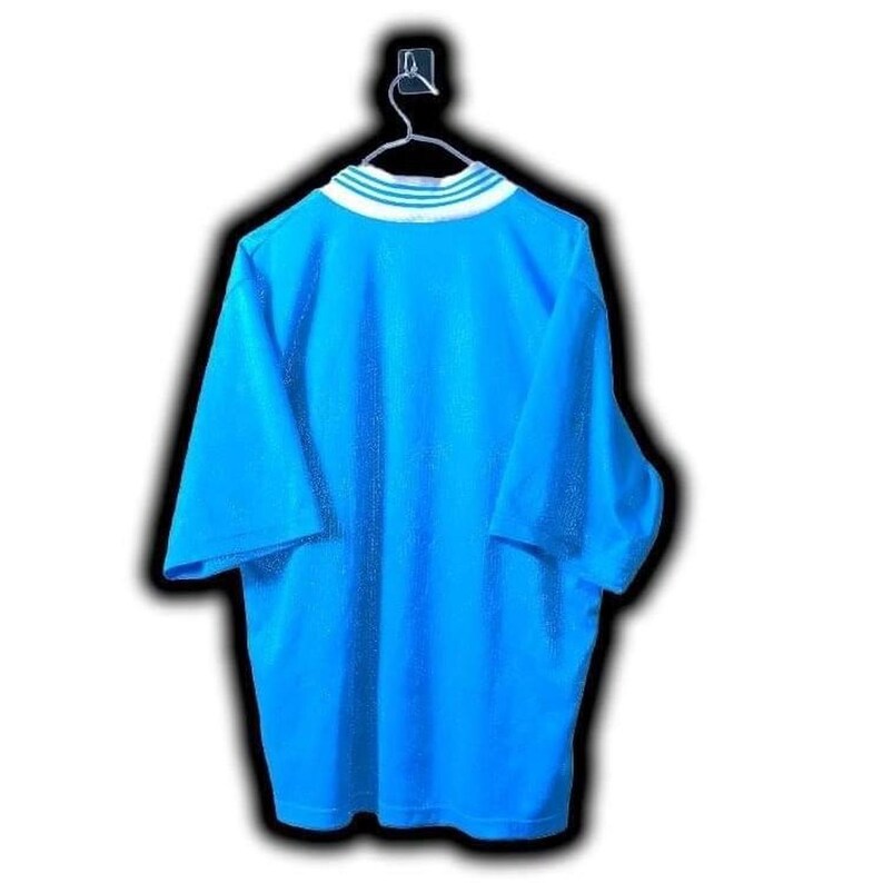 Puede incluir: Una camiseta azul claro de manga corta colgada de una percha. La camiseta tiene un cuello a rayas blancas y verdes. La camiseta est&aacute; hecha de un material suave y c&oacute;modo.