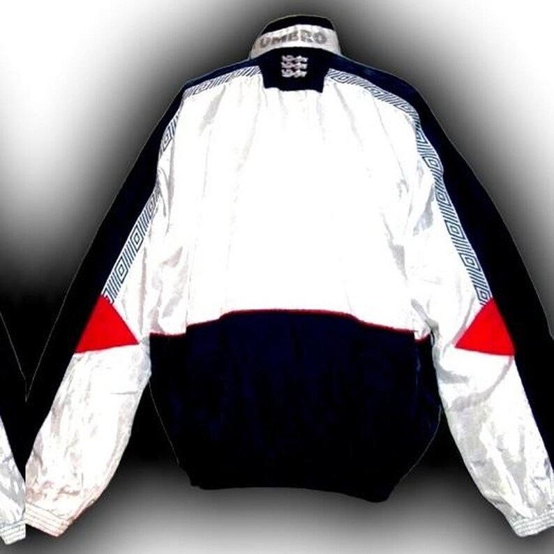 May include: A vintage Umbro England soccer jacket. The jacket features a white upper section, a navy blue lower section, and black sleeves. Red triangles accent the sides. The Umbro logo and England crest are visible on the collar.