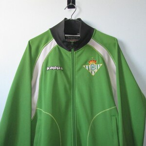 K&ouml;nnte beinhalten: Gr&uuml;ne Trainingsjacke mit Rei&szlig;verschluss und wei&szlig;en und schwarzen Akzenten. Die Jacke hat das Kappa-Logo und ein Team-Emblem. Die Jacke h&auml;ngt an einem Metallb&uuml;gel.
