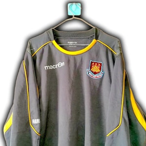 Puede incluir: Top deportivo gris de manga larga con ribete amarillo y el logo de Macron. Presenta el escudo del West Ham United en el pecho. La parte superior est&aacute; colgada de una percha.