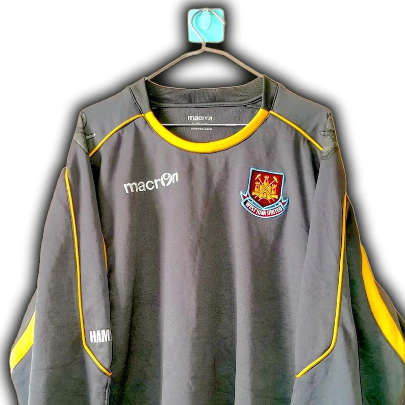 Puede incluir: Top deportivo gris de manga larga con ribete amarillo y el logo de Macron. Presenta el escudo del West Ham United en el pecho. La parte superior est&aacute; colgada de una percha.