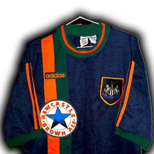 May include: Navy blue soccer jersey with orange and green accents. Features the Adidas logo, a Newcastle Brown Ale patch, and a team crest. The jersey is hanging on a white hanger.