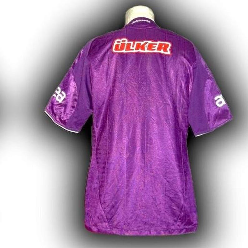 May include: A purple soccer jersey with the word "&Uuml;LKER" in red block letters across the back. The jersey has short sleeves with white trim and the letter "a" on the right sleeve. The jersey is made of a shiny, textured fabric.