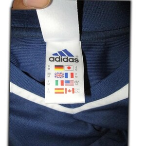May include: Close-up of a navy blue Adidas shirt tag. The tag features the Adidas logo and country flags, including the USA, Japan, and Canada. The shirt has white stripes and trim.