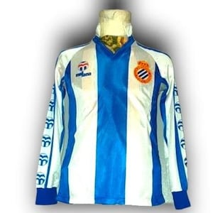Puede incluir: Una camiseta de fútbol de manga larga con un diseño de rayas blancas y azules. La camiseta presenta un cuello en V, puños azules y un escudo del equipo en el pecho. Las mangas tienen un patrón de logotipo repetido.