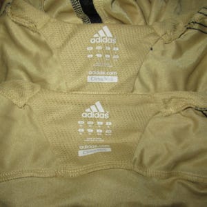 K&ouml;nnte beinhalten: Nahaufnahme von zwei beigefarbenen Sporthemden mit dem Adidas-Logo und Gr&ouml;&szlig;enetiketten. Die Hemden haben ein Mesh-Panel und den Text "adidas.com" und "Clima365".