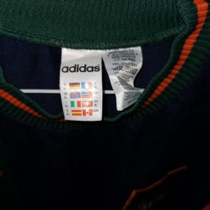 May include: Close-up of an Adidas label on a dark blue garment. The label features the Adidas logo, size indicators, and country flags. The garment has a green collar with orange stripes.