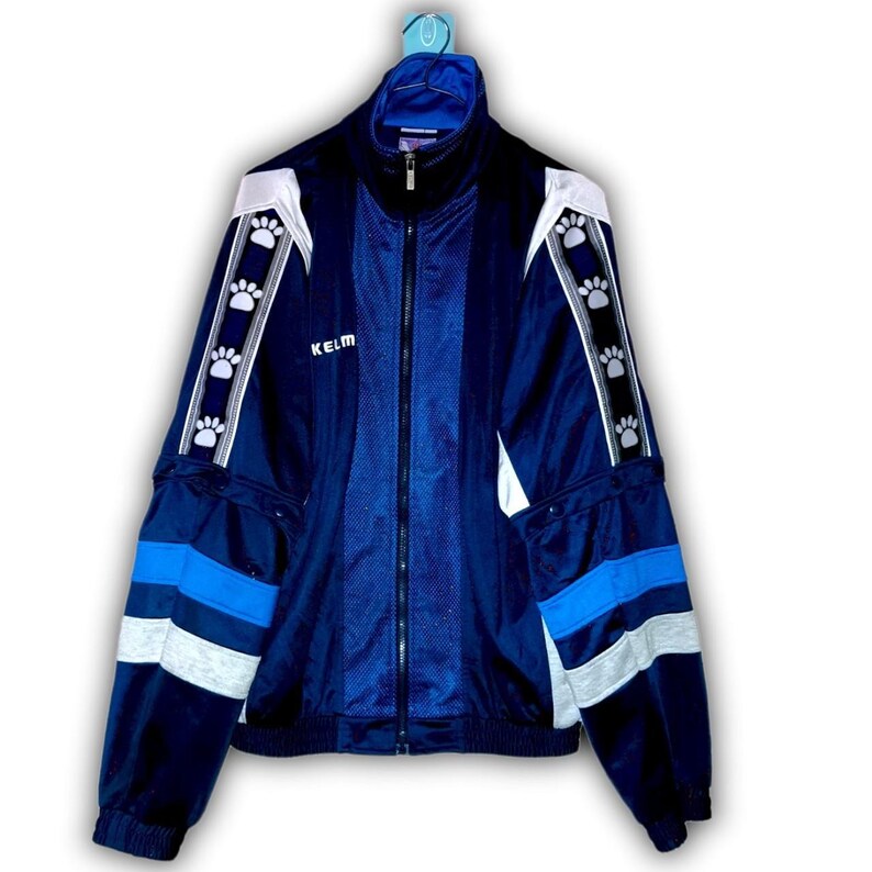 K&ouml;nnte beinhalten: Marineblaue Trainingsjacke mit wei&szlig;en und hellblauen Akzenten. Die Jacke hat einen durchgehenden Rei&szlig;verschluss, einen hohen Kragen und den Markennamen "KELM" auf der Brust. Pfotenabdruck-Designs zieren die &Auml;rmel.
