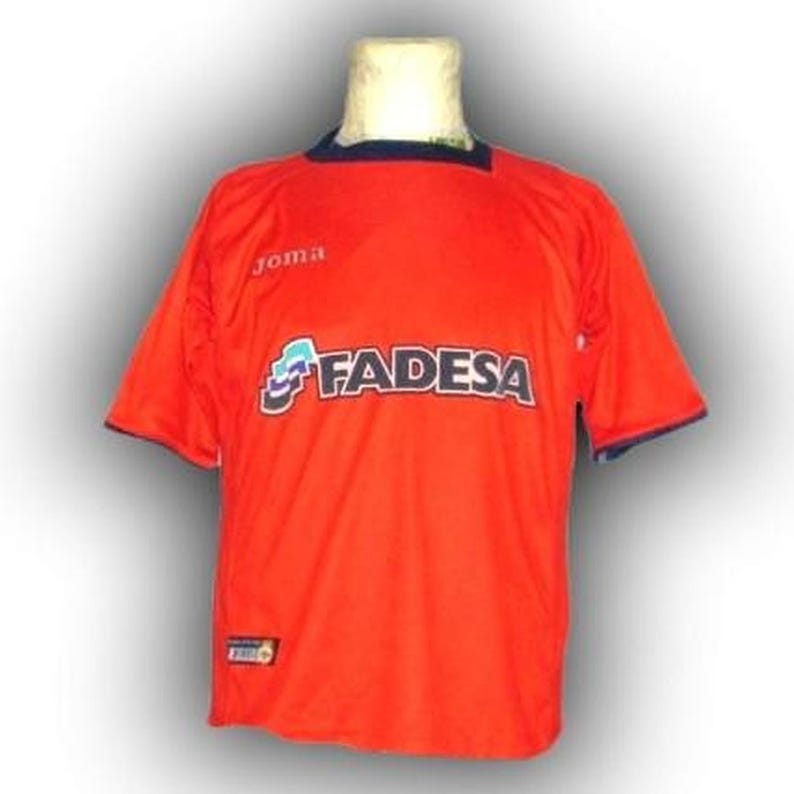 Op de afbeelding: Een rood voetbalshirt met korte mouwen met het woord "Joma" op de borst. Het woord "Fadesa" is in het midden gedrukt. Het shirt heeft marineblauwe afwerking op de kraag en mouwen. Een klein label is links onderaan zichtbaar.