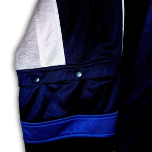 K&ouml;nnte beinhalten: Nahaufnahme eines marineblauen Jacken&auml;rmels mit einem wei&szlig;en und grauen Einsatz. Der &Auml;rmel hat zwei marineblaue Kn&ouml;pfe und einen blauen Streifen am B&uuml;ndchen. Die Jacke scheint aus einem glatten, m&ouml;glicherweise sportlichen Material zu sein.