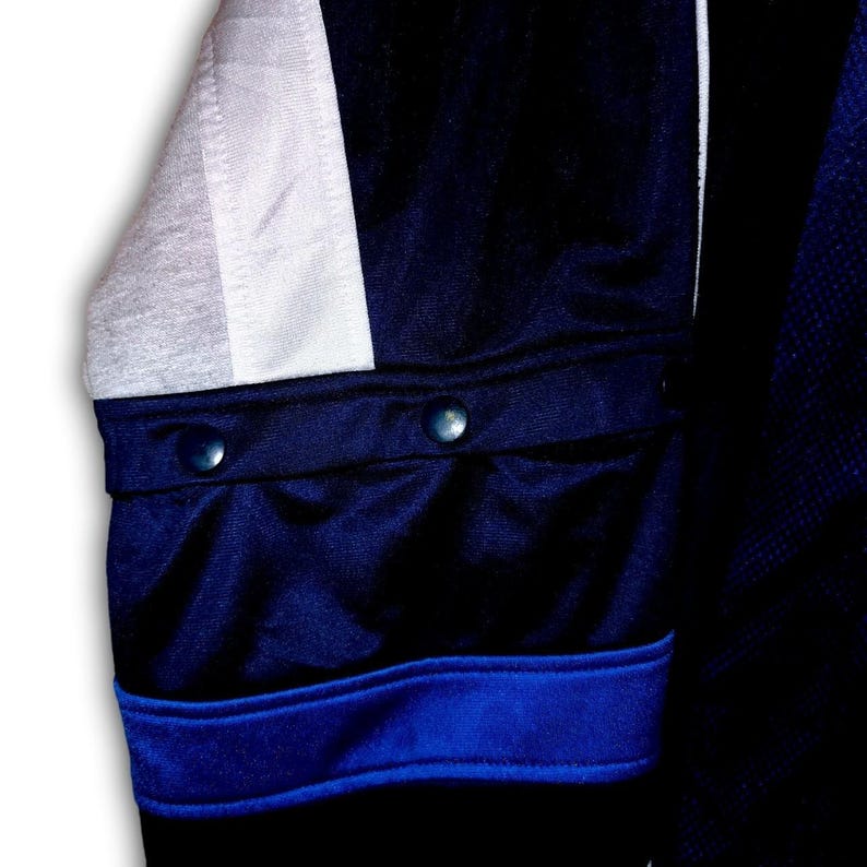 K&ouml;nnte beinhalten: Nahaufnahme eines marineblauen Jacken&auml;rmels mit einem wei&szlig;en und grauen Einsatz. Der &Auml;rmel hat zwei marineblaue Kn&ouml;pfe und einen blauen Streifen am B&uuml;ndchen. Die Jacke scheint aus einem glatten, m&ouml;glicherweise sportlichen Material zu sein.