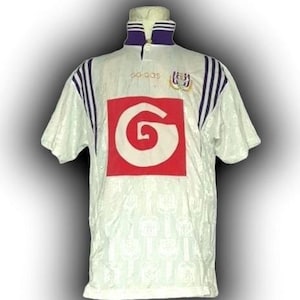 Puede incluir: Camiseta de fútbol blanca con detalles morados y azul marino. La camiseta presenta un gran cuadrado rojo con un diseño de remolino blanco, un cuello morado y el logotipo de Adidas. El escudo del equipo está en el pecho derecho.