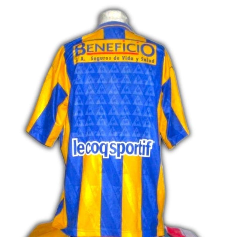 May include: A blue and yellow soccer jersey with the word "BENEFICIO" across the top and "le coq sportif" at the bottom. The jersey has blue and yellow vertical stripes and a blue patterned design.