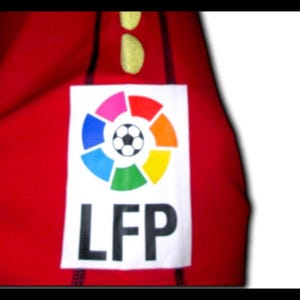 May include: Red fabric with a white rectangular patch featuring the LFP logo. The logo includes a colorful circular design with a soccer ball in the center. The letters "LFP" are printed in black below the logo.