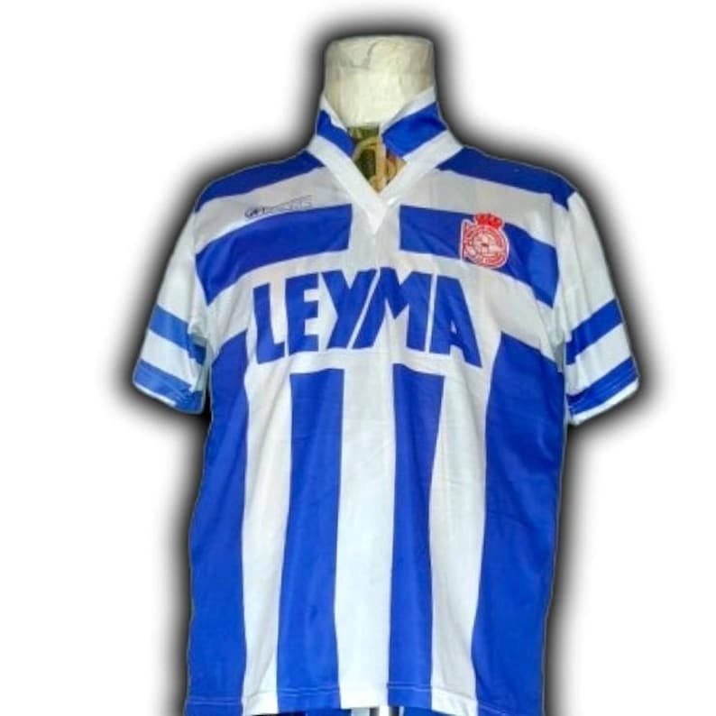 May include: A vintage soccer jersey with a blue and white striped design. The jersey features the word "LEYMA" in large blue letters across the chest. A team logo is visible on the upper right chest area. The collar and sleeves have blue accents.